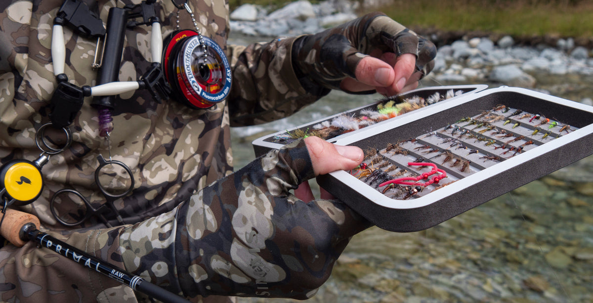 Angler holding an open fly box filled with trout flies beside a rocky river, with a Primal fly rod tucked under their arm